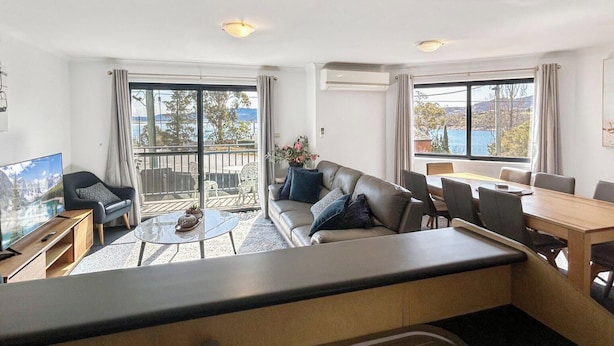 Una sala de estar moderna con sofá, mesa de centro y televisor, ventanas amplias con vistas, y un balcón con mobiliario exterior.