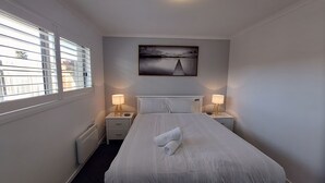 Una habitación de hotel con cama, dos mesitas de noche con lámparas, una ventana con persianas y un cuadro en la pared.