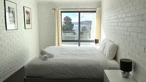 A hotel room with a large bed, a nightstand, a lamp, and a window with a view of the outdoors. - The Summit 15 - Spacious 4 bedroom with lake views (Jindabyne)