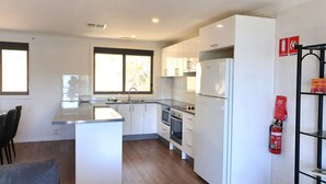 A modern kitchen with white cabinets, a built-in oven, and a refrigerator. There is a window providing natural light and a fire extinguisher mounted on the wall. - The Summit 15 - Spacious 4 bedroom with lake views (Jindabyne)