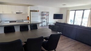 A modern kitchen with white cabinets, a dining area with a table and chairs, and a living area with a sofa and a TV. - The Summit 15 - Spacious 4 bedroom with lake views (Jindabyne)