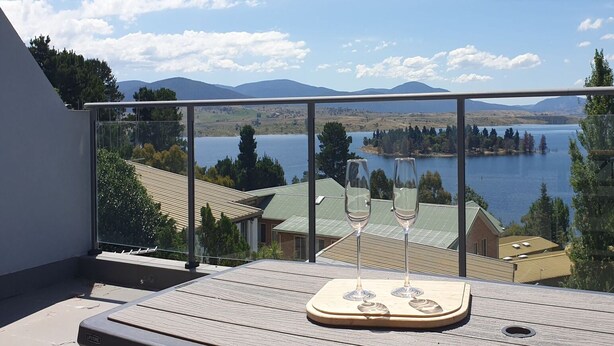 A balcony with a wooden deck, a glass railing, and a view of a lake and mountains. - The Summit 15 - Spacious 4 bedroom with lake views (Jindabyne)