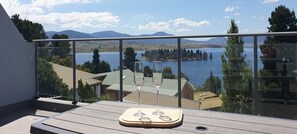 Un balcón con cubierta de madera, barandilla de vidrio y vista a un lago y montañas.