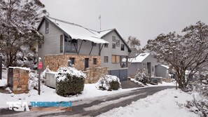 A two-story house with a stone foundation and grey siding, surrounded by snow-covered trees and a snow-covered driveway. - Ramshead Creek 1 - Crackenback Ridge (Crackenback Ridge)