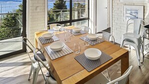 A dining area with a wooden table set for four, white chairs, and a view of the sea through large windows. - The Summit 16 - Huge deck with lake views (Jindabyne)