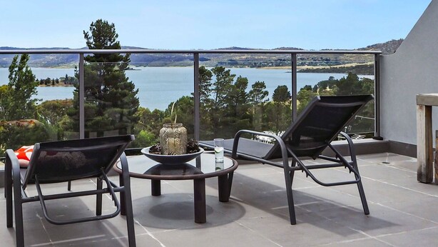 A balcony with two black chairs, a small round table, and a decorative vase. The balcony overlooks a lake with trees and distant hills. - The Summit 16 - Huge deck with lake views (Jindabyne)