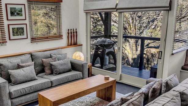 Een woonkamer met een grijze bank, een houten salontafel en een balkon met een barbecue en tuinmeubilair.