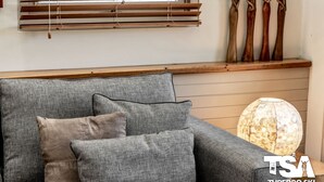 Een grijze bank met bijpassende kussens, een houten plank met decoratieve objecten, en een lamp met een bijzondere textuur.