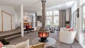 A modern living room with a central wood-burning stove, a staircase, a dining area, and large windows. - Rileys Run - Woodridge (Woodridge)