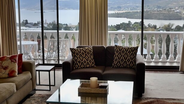 A living room with a sofa and armchairs, a coffee table, and a view of a lake and mountains through large windows.