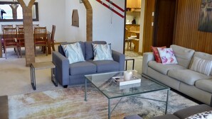 A living room with a grey sofa, a glass coffee table, and a beige sofa with floral cushions. There is a dining area with wooden chairs and a table in the background.