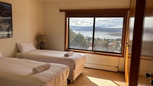A hotel room with two beds, a window with a view of a lake and town, and a framed picture on the wall.