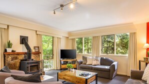 Una sala de estar acogedora con chimenea, asientos cómodos, televisor y ventanas amplias con vistas a la vegetación.