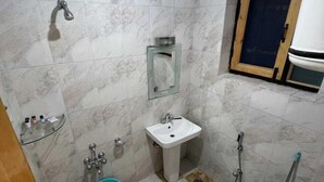 A small bathroom with a toilet, sink, and shower. There is a mirror above the sink and a window with a wooden frame. - HOTEL WEST INN BOULEVARD (Srinagar)