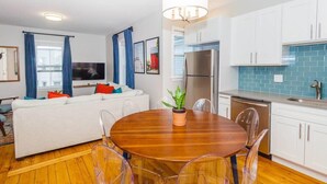 A modern living room with a white sofa, a wooden dining table with clear chairs, a kitchen with white cabinets and blue backsplash, and a large window with blue curtains. - Cute Boston Condo | Near T (Boston)