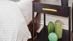 A neatly made bed with a grey and white patterned bedspread, a wooden headboard, a bedside table with a lamp, a vase with white flowers, and two green cacti. - Cute Boston Condo | Near T (Boston)