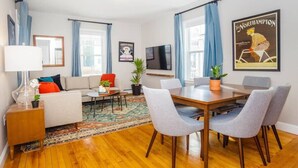 A modern living room with a sofa, a coffee table, and a dining table with chairs. There is a TV mounted on the wall and a poster on the wall. - Cute Boston Condo | Near T (Boston)