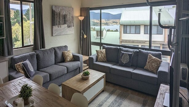 A modern living room with grey sofas, a wooden coffee table, and a view of a lake and buildings. - Alpine Mountain View 40 (Jindabyne)