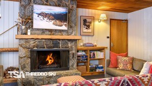 Uma sala de estar aconchegante com lareira de pedra, televisão na parede, estante de livros e um espaço de estar confortável.