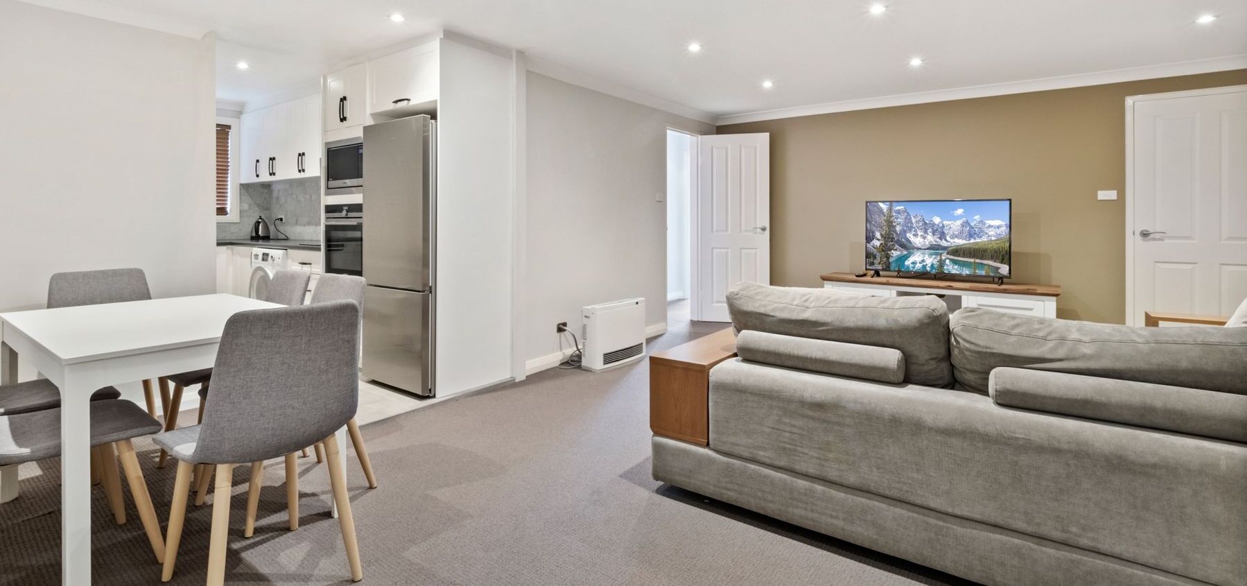 Un salon moderne avec un canapé gris, une table à manger avec des chaises, un coin cuisine équipé d’appareils électroménagers et un téléviseur posé sur un meuble en bois.