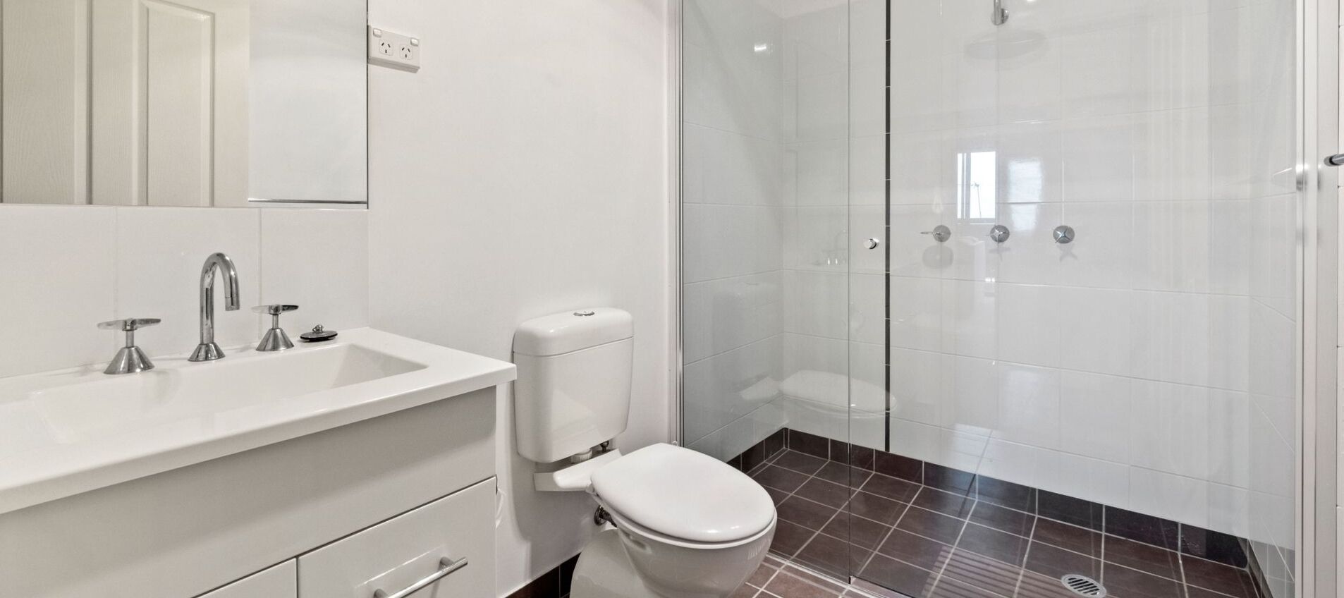 A modern bathroom with a white sink, three faucets, a toilet, and a glass-enclosed shower area.