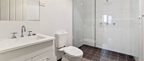 A modern bathroom with a white sink, three faucets, a toilet, and a glass-enclosed shower area.