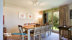 A dining area with a wooden table and chairs, framed artwork on the wall, and a view of greenery outside.