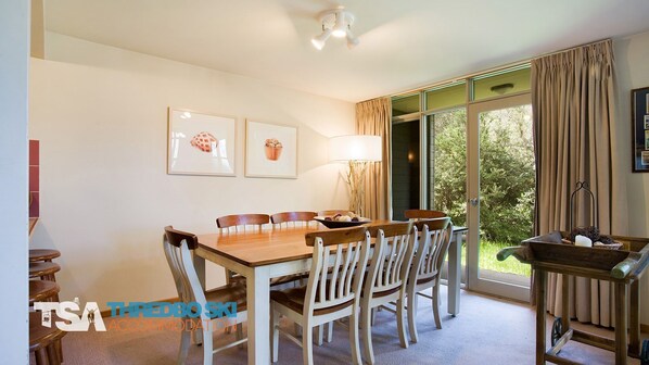 A dining area with a wooden table and chairs, framed artwork on the wall, and a view of greenery outside.