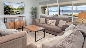 A living room with a sofa, armchairs, a coffee table, a TV, a wooden cabinet, and a large window with a view of a lake and mountains. - Birralee 1 great lake and mountain views (Jindabyne)