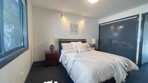 Un dormitorio con una cama, mesitas de noche, una ventana y una puerta.