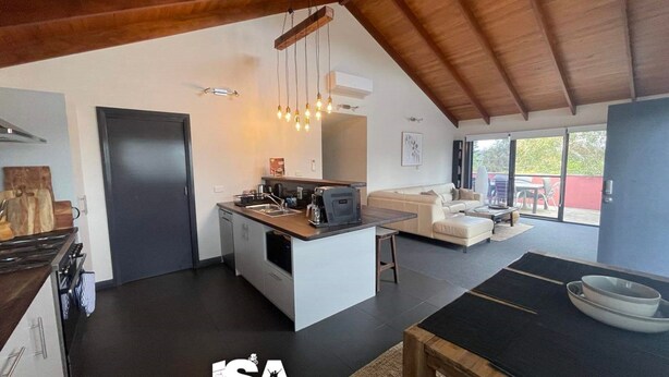 Una cocina moderna con una isla central, un comedor con sofá y un amplio ventanal corredizo que da a un patio exterior.