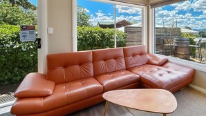 Un rincón amoblado con un sofá de cuero y una mesita de madera, ventanas amplias que ofrecen vista a un patio trasero con cercas, árboles y una cadena montañosa distante.