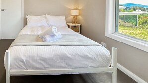 Una cama bien hecha con sábanas blancas y una toalla enrollada, una lámpara de noche y una ventana con vistas a zonas verdes y edificios.