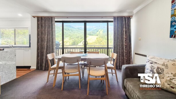 A modern living room with a dining area, large windows with curtains, a sofa, and a kitchen area. - Mowamba D4 - Central Village (Central Village)