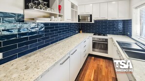 A modern kitchen with white cabinets, a blue tiled backsplash, a built-in microwave, and a stainless steel oven. - Mowamba D4 - Central Village (Central Village)