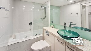 A modern bathroom with a glass-enclosed shower, a white toilet, and a large mirror with a green glass sink. - Mowamba D4 - Central Village (Central Village)