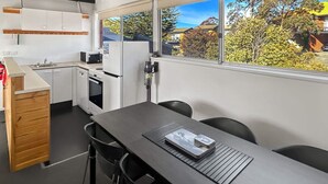 Une cuisine compacte avec des meubles blancs, une cuisinière et un évier. On y trouve une table à manger avec des chaises et une fenêtre donnant sur des arbres et des maisons.