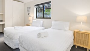 A hotel room with two single beds, a window with a view of greenery, a bedside table with a lamp, and a wardrobe.