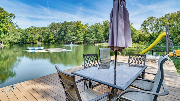 A wooden deck with a glass table and chairs, overlooking a calm lake with a dock and a yellow slide. - Lake Escape w/ Hot Tub & Docks in Hope! (Hope)
