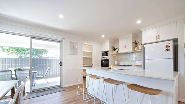Een moderne keuken met witte kasten, een centraal eiland met barstoelen, een koelkast en een schuifdeur naar een buitenruimte.
