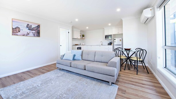 A modern living room with a grey sofa, a dining table with chairs, and a kitchen area in the background. - Cozy Getaway - Modern Central Unit | Garden View (Dubbo)