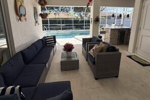 A poolside seating area with two sofas and a coffee table, a view of a swimming pool, and a patio with a stone fireplace. - Cape Coral Pool Home w/ Outdoor Kitchen & Games (Cape Coral)