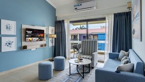 A modern living room with a blue sofa, a round coffee table with a bowl of fruit and glasses, a TV displaying 'Stranger Things', and a balcony with a view of a building. - 1BR w Pool Sauna Near Calangute Baga Beach (Calangute)