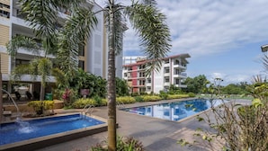 A hotel pool area with two pools, palm trees, and a building with balconies in the background. - 1BR w Pool Sauna Near Calangute Baga Beach (Calangute)