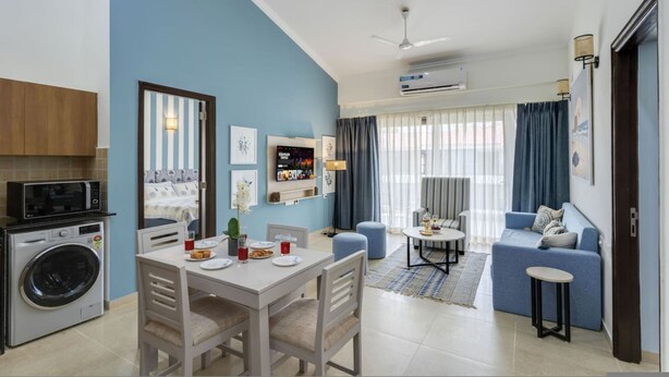 A modern living room with a sofa, a dining table with chairs, a kitchen area with a washing machine, and a TV on the wall. - 1BR w Pool Sauna Near Calangute Baga Beach (Calangute)