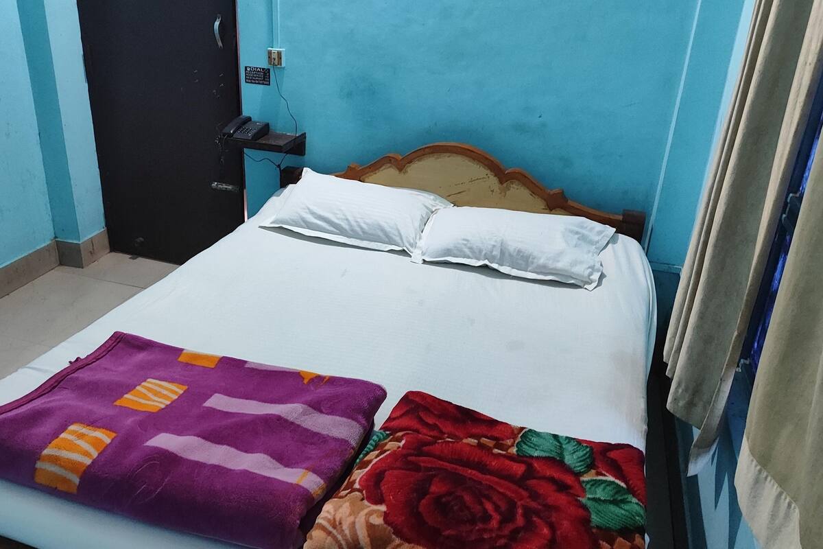 Una cama individual con sábanas y almohadas blancas, una colcha morada y naranja, y una manta floral roja. A la izquierda de la cama hay una silla roja. La habitación tiene paredes azules y un cabecero de madera.