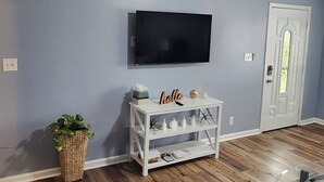 A room with a flat-screen TV mounted on the wall, a white console table with decorative items, a potted plant, and a wooden floor. - Senseney Cottage~Pet Welcome, Fenced Yard (Clarksville)