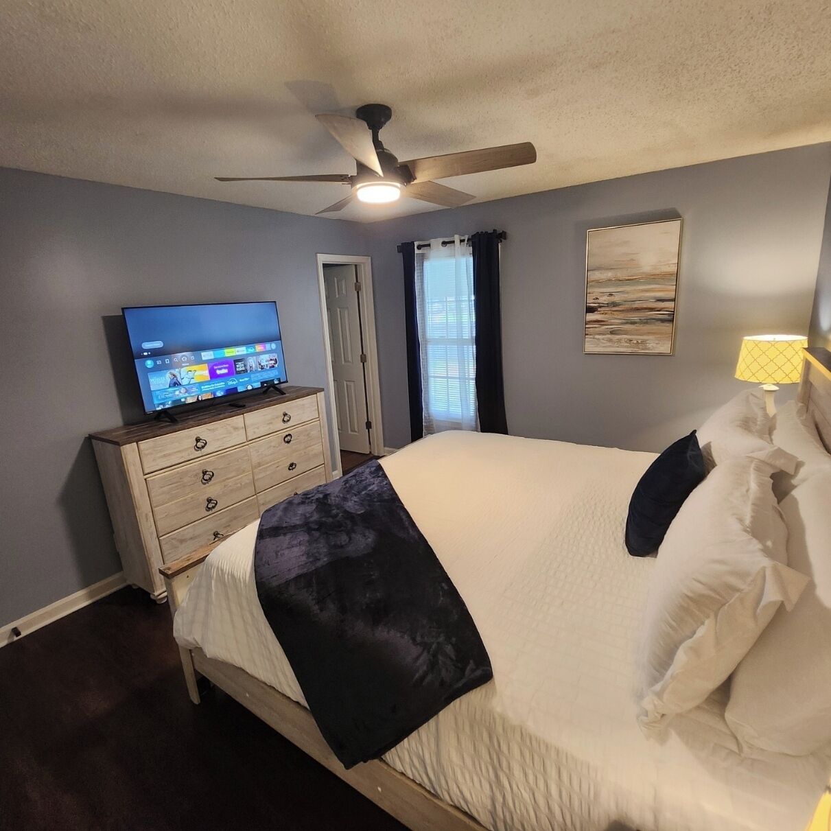 Ein Schlafzimmer mit einem Bett, einem Fernseher auf einer Kommode, einem Deckenventilator und einem Fenster mit Vorhängen.