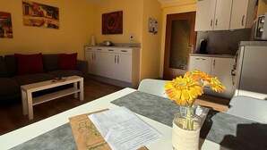 Een knus woongedeelte met een bank, een eettafel met een bloevas, een keuken met kastjes, en kunstwerken aan de wanden.