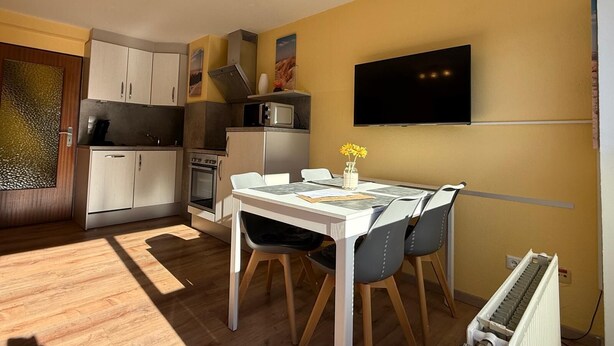 Een kleine keuken met witte kasten, een magnetron en een fornuis. Er staat een eettafel met stoelen en een vaas met bloemen. Een flatscreen televisie is aan de muur bevestigd.